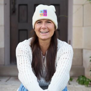 A person smiling and wearing the Moose Tracks tan beanie with pop art patch on the front.