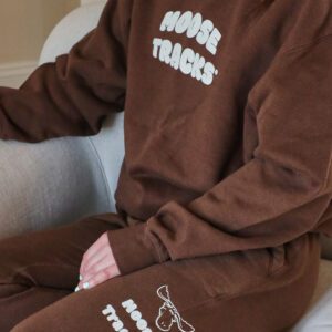 A person wearing a matching brown sweatsuit, sitting on a sofa. The crewneck reads "MOOSE TRACKS" in white letters on the chest and the pants have a white moose head logo and "MOOSE TRACKS" printed on the side.