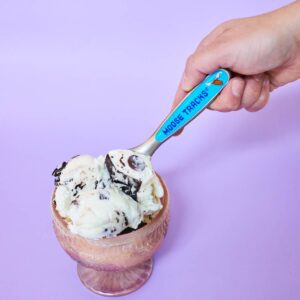 A hand holding the Moose Tracks Perfectly Pint-Sized Ice Cream Scoop next to a bowl of ice cream. The scoop has a blue design, moose head logo and reads, "Moose Tracks" on the handle.