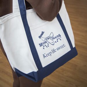 Someone carrying the Moose Tracks "Keep Life Sweet" canvas tote bag. Bag has navy detailing and a Moose Tracks logo and text reading "Keep Life Sweet" in navy-colored screen printing.