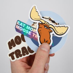 A hand holding up an ensemble of Moose Tracks stickers, with a moose icon sticker on top