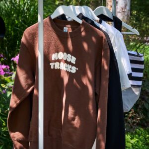 A brown crewneck sweatshirt that reads, "Moose Tracks" in vanilla-colored screen printing hanging on a clothes rack.