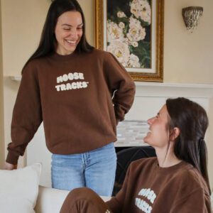 Two women are wearing matching brown sweatshirts with white text reading "MOOSE TRACKS" on the front. One woman is standing and smiling at the other, who is seated and looking up.