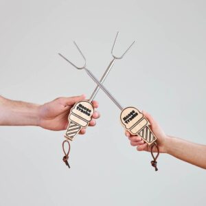 Two hands holding two unextended Moose Tracks S'mores Sticks on either side of the frame. Chrome-plated s’mores sticks with light tan wooden handles shaped like ice cream cones. The handles are engraved with "Moose Tracks" in black lettering and finished with a leather loop at the base.