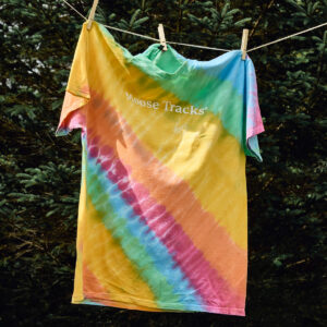 Moose Tracks Tie-Dye T-Shirt hanging on a clothesline with trees in the background. The shirt features a rainbow pastel tie-dye pattern with white screen-printed text in the center that reads "Moose Tracks".
