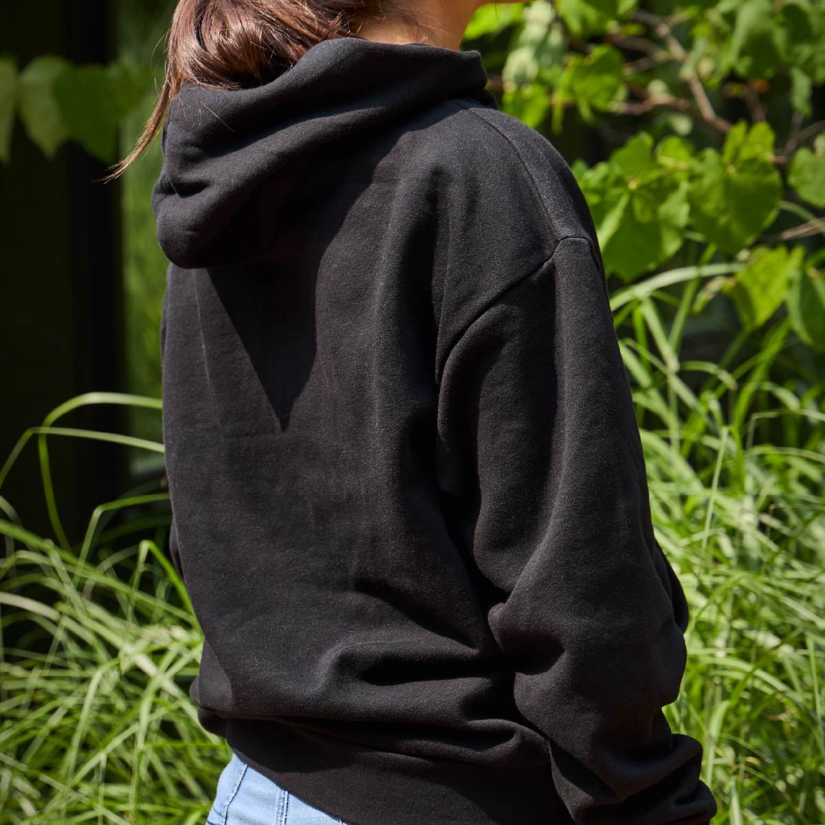A woman wearing the Moose Tracks Keep Life Sweet Hoodie in front of trees and green bushes. Image shows the back of the sweatshirt, which is black and has a hoodie.