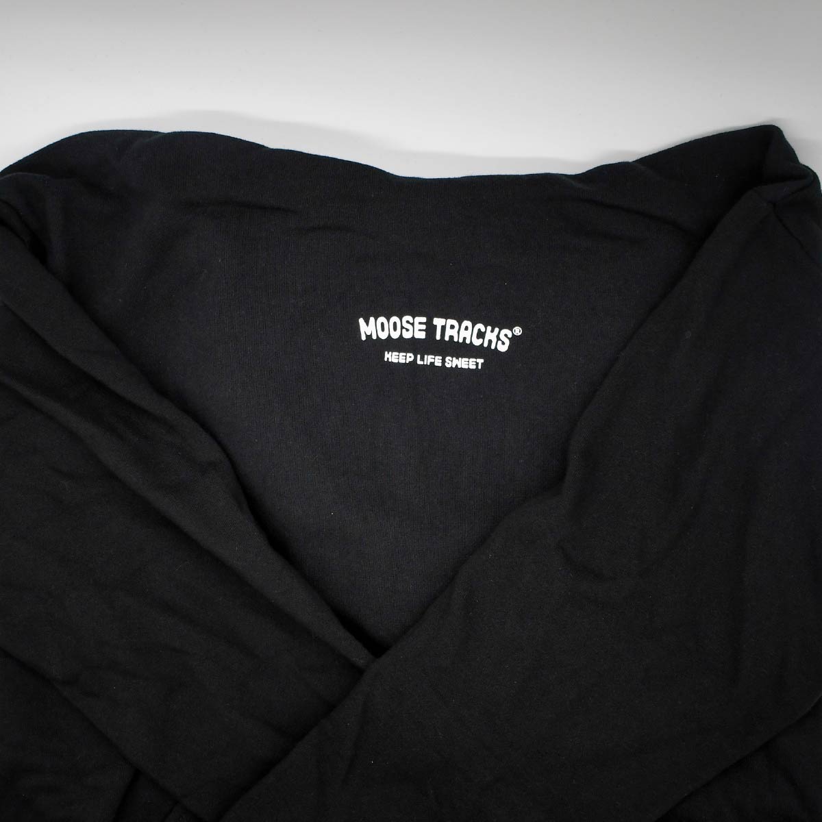 A close-up image of the front of the Moose Tracks Keep Life Sweet Hoodie. The sweatshirt is black with the words "Moose Tracks" and "Keep Life Sweet" in screen-printed white text on the chest. The hoodie is tucked behind the sweatshirt and the sleeves are folded in front of it.