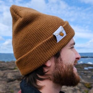 A man with a beard wearing a brown Moose Tracks rib-knit cuffed beanie against a backdrop of a lake and blue sky. The beanie is cuffed and features a moose head icon on a white label.